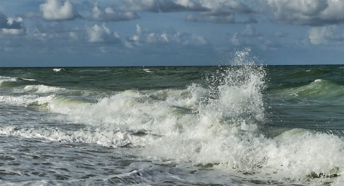 A wave crashes and splashes against the shore under a cloudy sky.