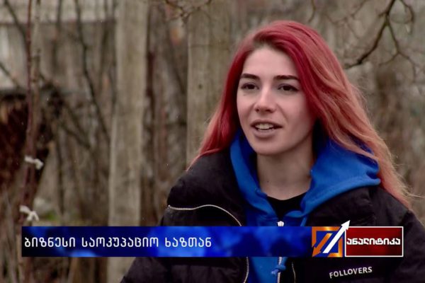 A woman with red hair wearing a blue and black jacket speaks outdoors with a blurred forest background.