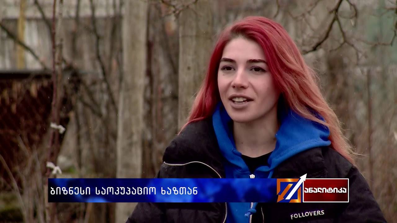 A woman with red hair wearing a blue and black jacket speaks outdoors with a blurred forest background.