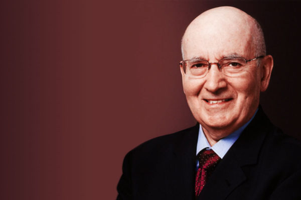 An elderly man with glasses, wearing a suit and tie, smiles warmly against a red background.