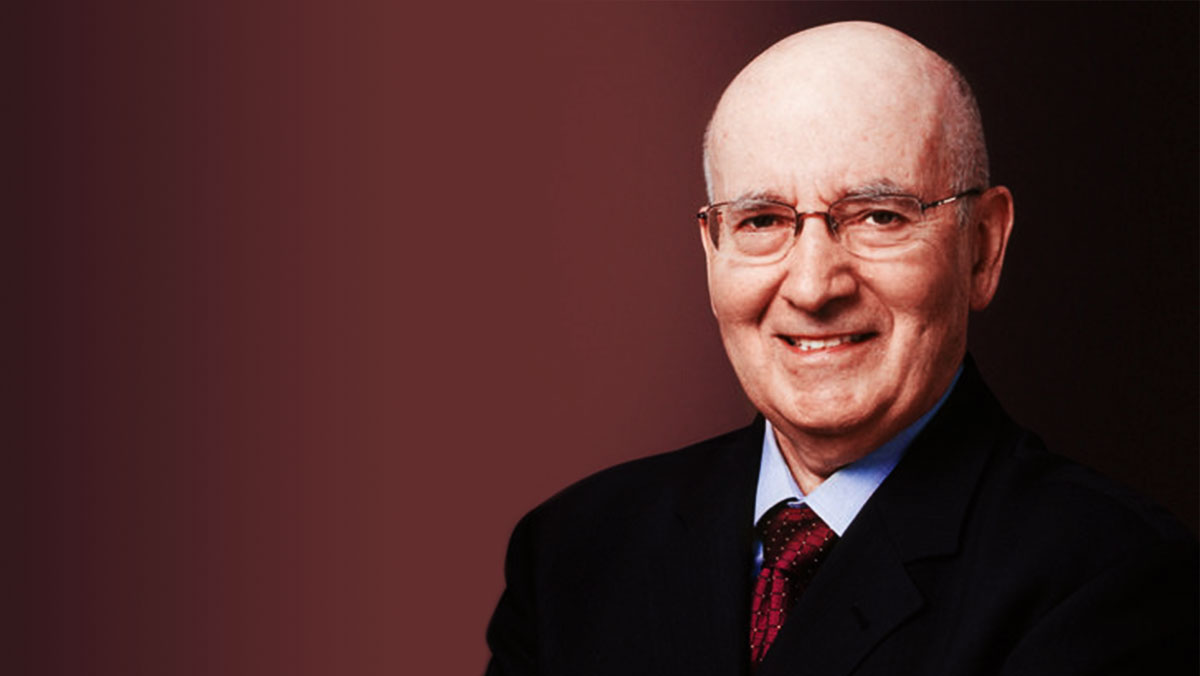 An elderly man with glasses, wearing a suit and tie, smiles warmly against a red background.