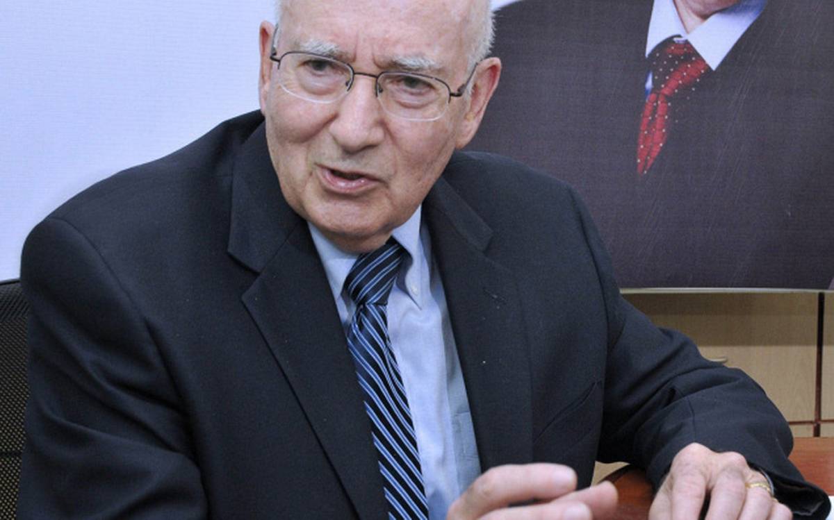 An elderly man in a suit and glasses gestures while speaking, seated in a formal setting.