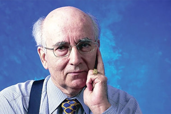 An elderly man wearing glasses and a striped shirt talks on a ring-shaped device held to his ear.