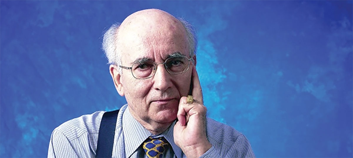 An elderly man wearing glasses and a striped shirt talks on a ring-shaped device held to his ear.