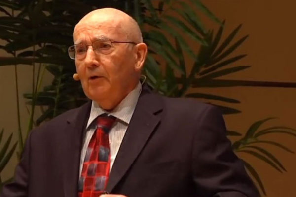 An elderly man wearing glasses, a suit, and a red tie speaks at a podium with plants in the background.
