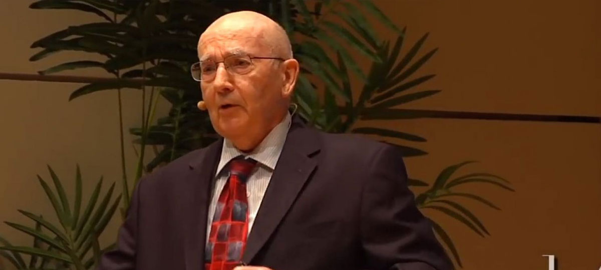 An elderly man wearing glasses, a suit, and a red tie speaks at a podium with plants in the background.