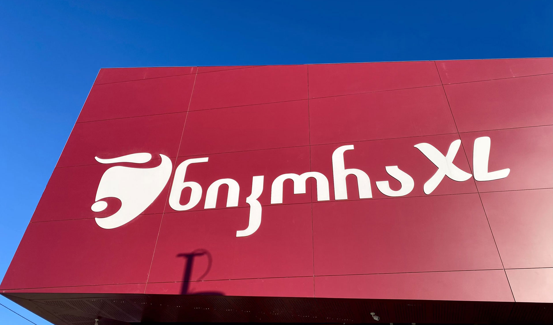 A red sign displays the text "Bigmhs XL" in white cursive letters against a clear blue sky.