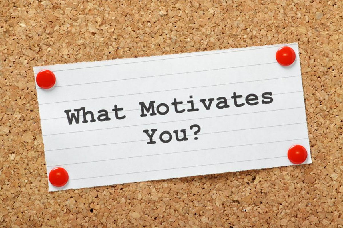 A note pinned to a corkboard asking, "What Motivates You?" with red pushpins.